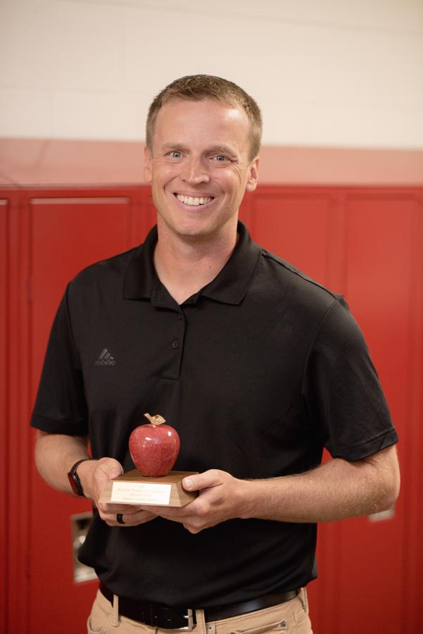 斯科特·菲利普斯(Scott Phillips)获得了2024年内布拉斯加州年度教师奖. 他身穿黑色polo衫，手持奖杯, 木底座上的红色花岗岩苹果, 就在他肚子前面. 他身后是奥罗拉中学的红色储物柜. 