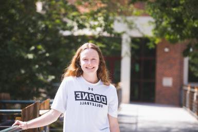 小麦肯齐·佩因特穿着一件白色t恤，上面写着“Doane Volleyball”.她站在谎言科学与数学大楼前.